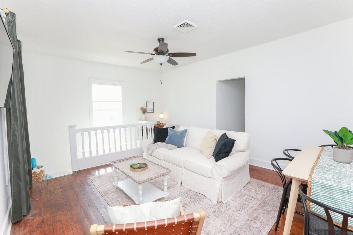 Uma sala de estar espaçosa com um sofá branco, uma mesa de centro, uma mesa de jantar com cadeiras, um ventilador de teto e uma varanda com guarda-corrimão.