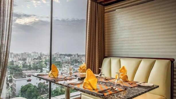 A dining area with a table set for four, a cushioned bench, and a city view through large windows. - Grand Palace Sylhet (Sylhet)
