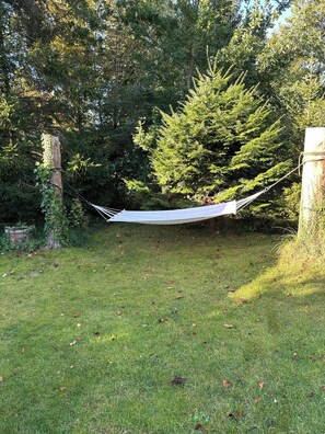 Un hamac est suspendu entre deux arbres, dans une zone herbeuse, avec un feuillage dense en arrière-plan.