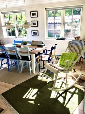 Un coin repas comprenant une table, des chaises et un fauteuil à bascule. Des fenêtres offrent une vue sur la verdure environnante.