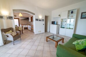 Una sala de estar con un sofá verde, un sillón de mimbre, una mesa de centro de vidrio y una cocina al fondo.