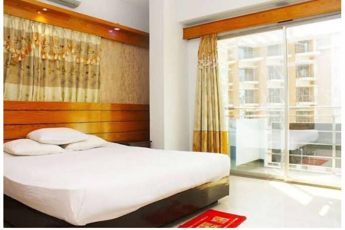 Quarto de hotel com cama, cabeceira de madeira e uma grande janela com cortinas.