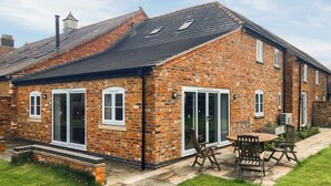 Une maison en briques avec un toit en tuiles, de grandes portes-fenêtres coulissantes et un patio aménagé avec du mobilier d’extérieur.