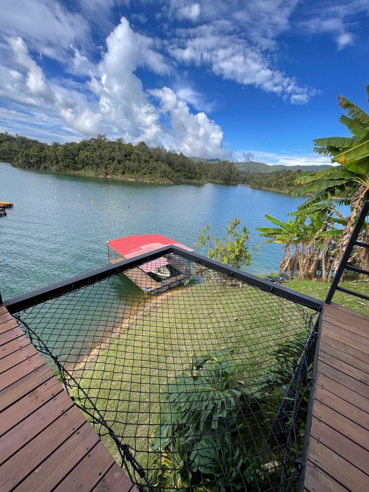 Eine Holzterrasse mit einem Netzbereich, der auf einen ruhigen See blickt, umgeben von üppiger Vegetation und blauem Himmel.