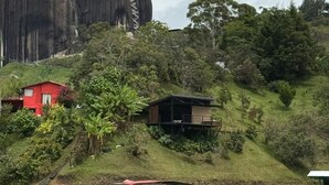 A coastal landscape with a prominent rock formation, a red building, and a small wooden structure on a hill. - Glamping Boreal (Guatapé)