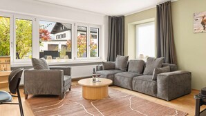 Una sala de estar moderna con un sofá gris, una mesa de centro redonda y vistas a una casa a través de grandes ventanales.