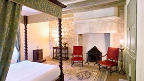 Een kamer met een hemelbed, twee rode fauteuils, een stenen openhaard en een kamerbreed tapijt met een patroon.