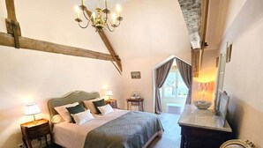 Un dormitorio con cama, mesitas de noche, escritorio, silla, candelabro y ventana con cortinas.
