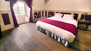 Un dormitorio con una cama grande, suelo de madera, una ventana con cortinas, una mesita de noche y una lámpara.
