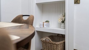 Un coin repas avec une table et des chaises en bois, une étagère blanche contenant des livres et un panier, et un interrupteur fixé au mur.