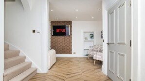 Un couloir moderne avec un parquet en chevrons, un escalier, un téléviseur fixé au mur qui affiche le logo « NETFLIX », et un fauteuil à motifs.