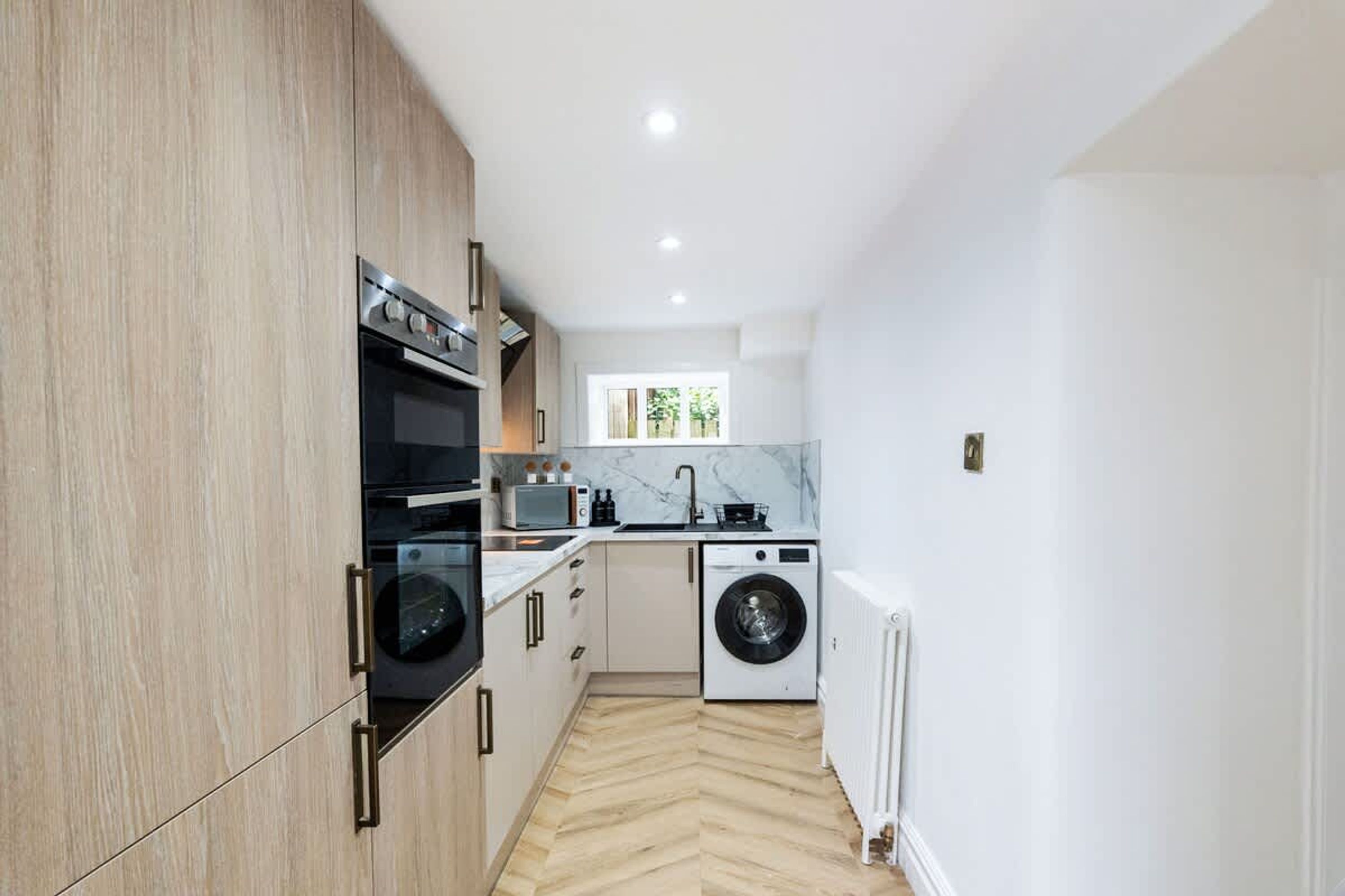 Cocina compacta con horno integrado, microondas y lavadora, armarios de madera y suelo con estampado en espiga.