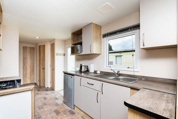 A compact kitchen with a dishwasher, a microwave, and a window above the sink. - 3 Bedroom Caravan - Seton Sands (Prestonpans)