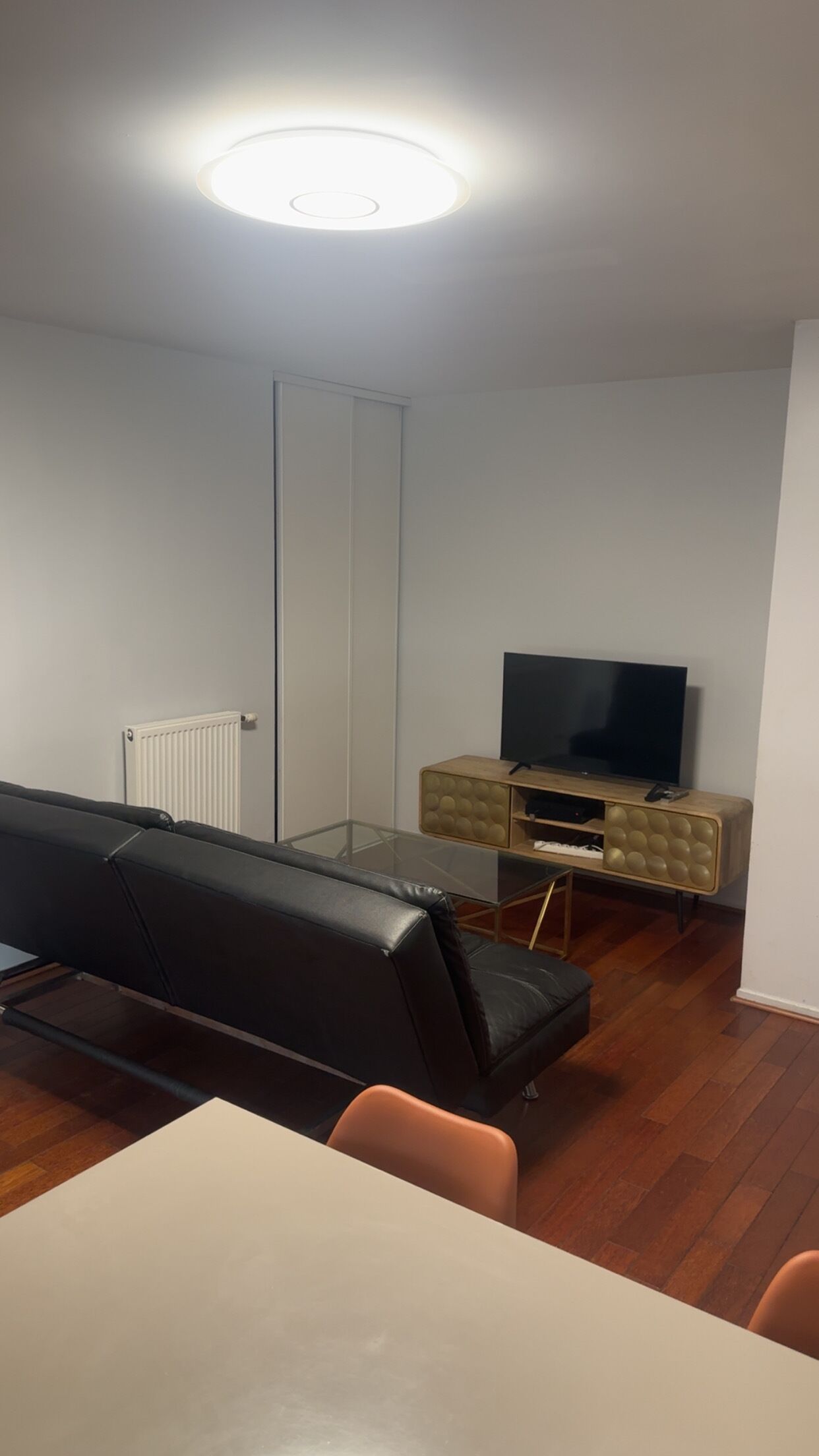 A modern living room with a sofa, a TV on a wooden cabinet, a glass coffee table, and a radiator.