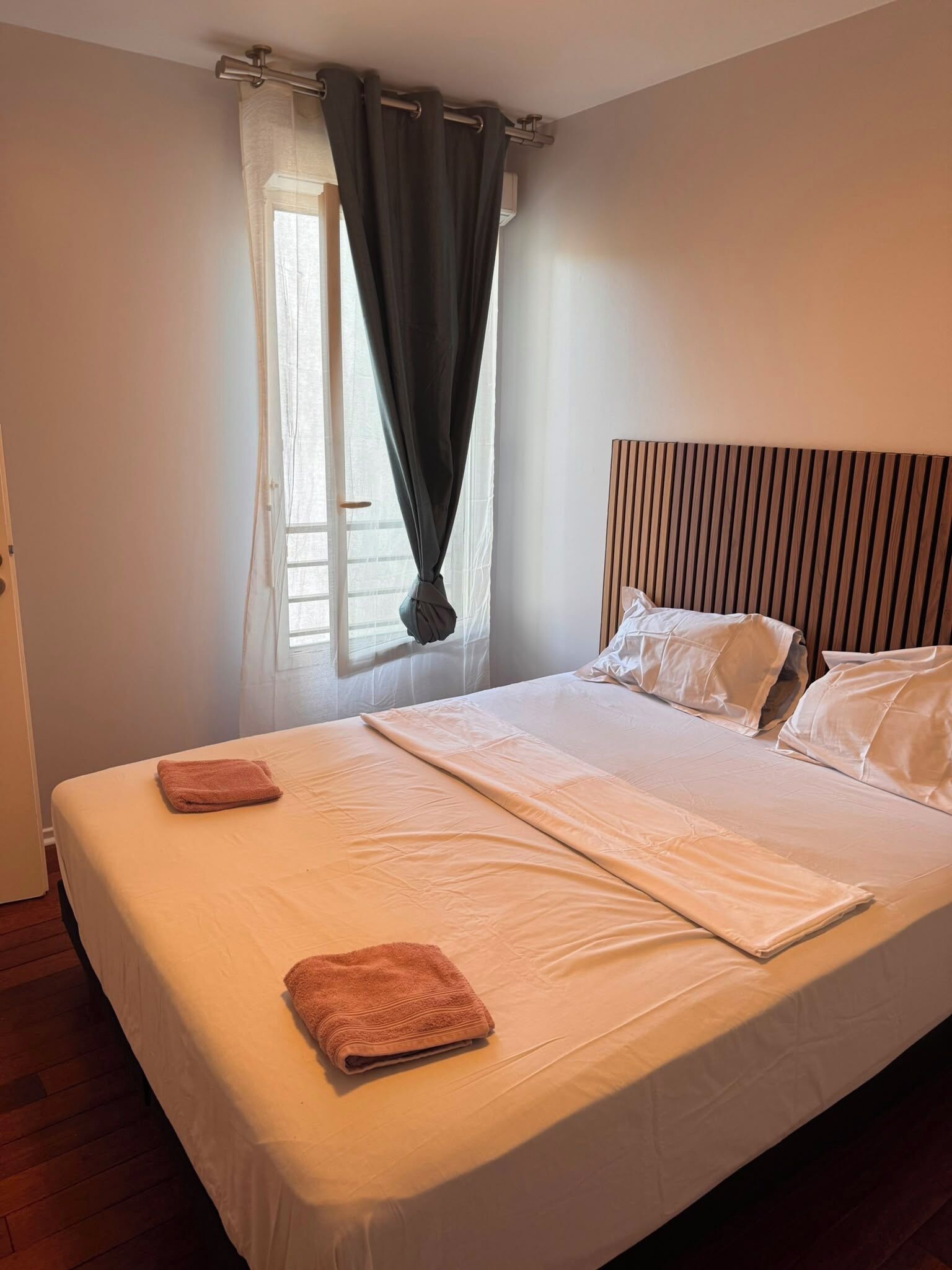 A neatly made bed with pillows and a folded towel, a window with curtains, and a wooden headboard.
