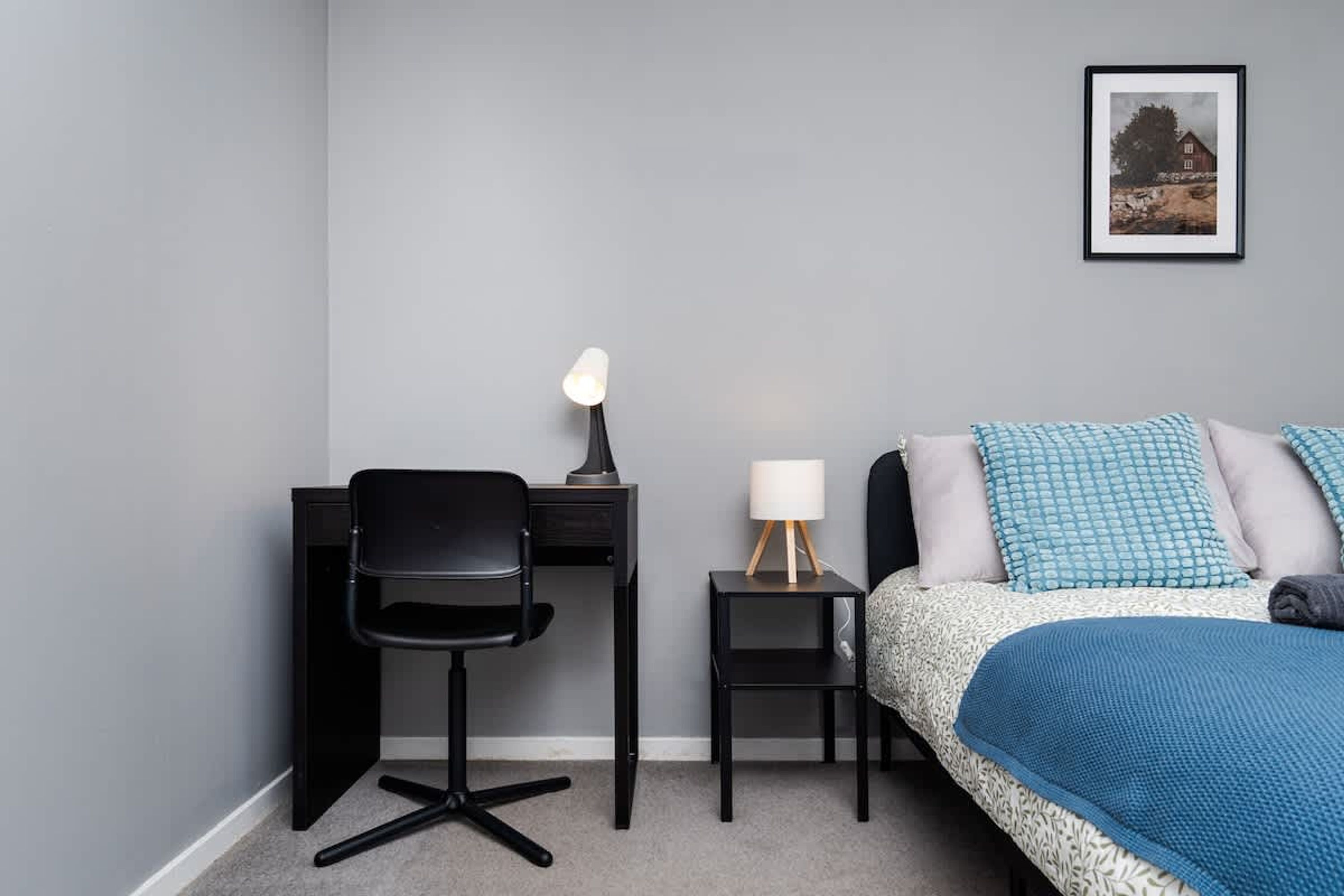 Ein Schlafzimmer mit einem Bett, einem Schreibtisch, einem Sessel, einer Lampe und einem gerahmten Bild an der Wand.