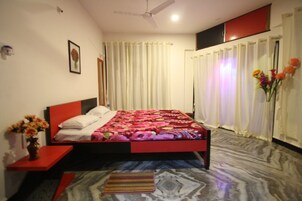 Ein Schlafzimmer mit einem Bett, einem Nachttisch, einer Vase mit Blumen, einem Deckenventilator und Vorhängen.