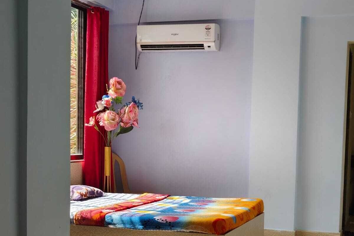 A bedroom with a bed, an air conditioner, a window with red curtains, and a vase with flowers.
