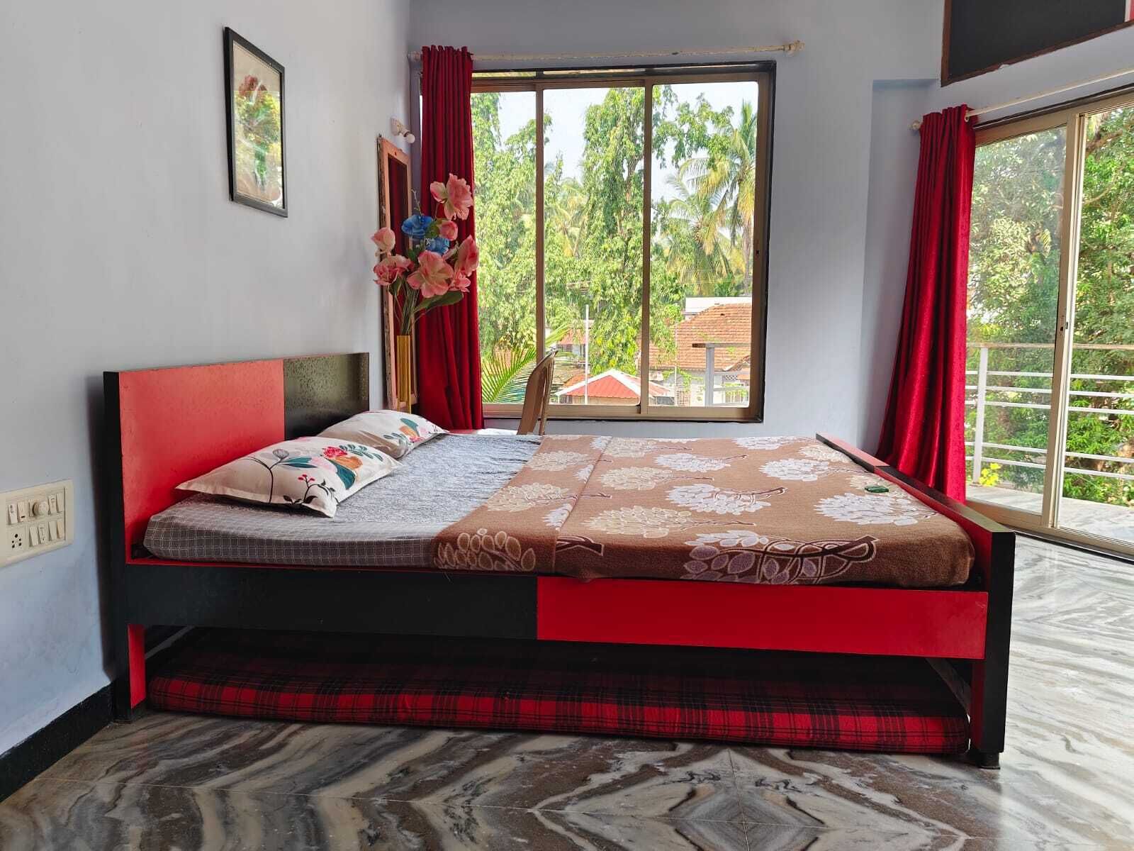 A bedroom with a bed, a window with red curtains, and a view of greenery outside.