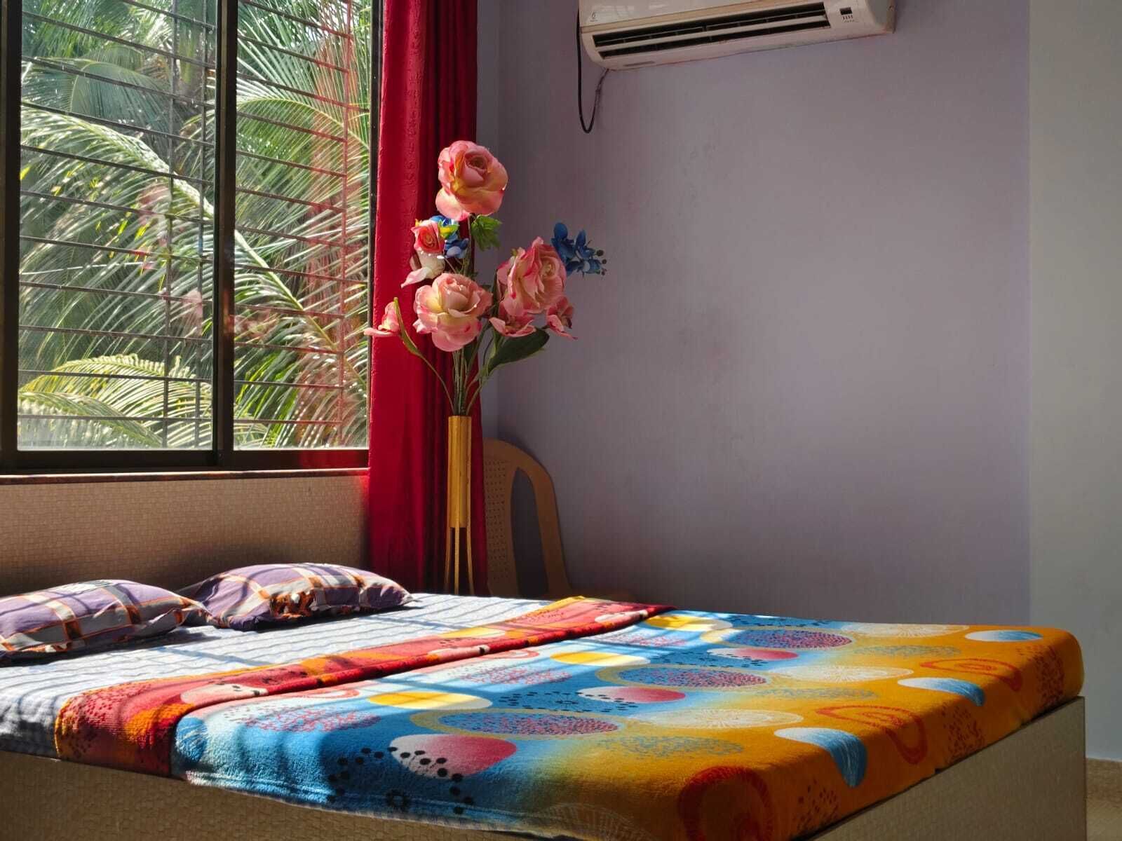 A bedroom with a bed, a window with a view of palm trees, a red curtain, and an air conditioning unit on the wall.