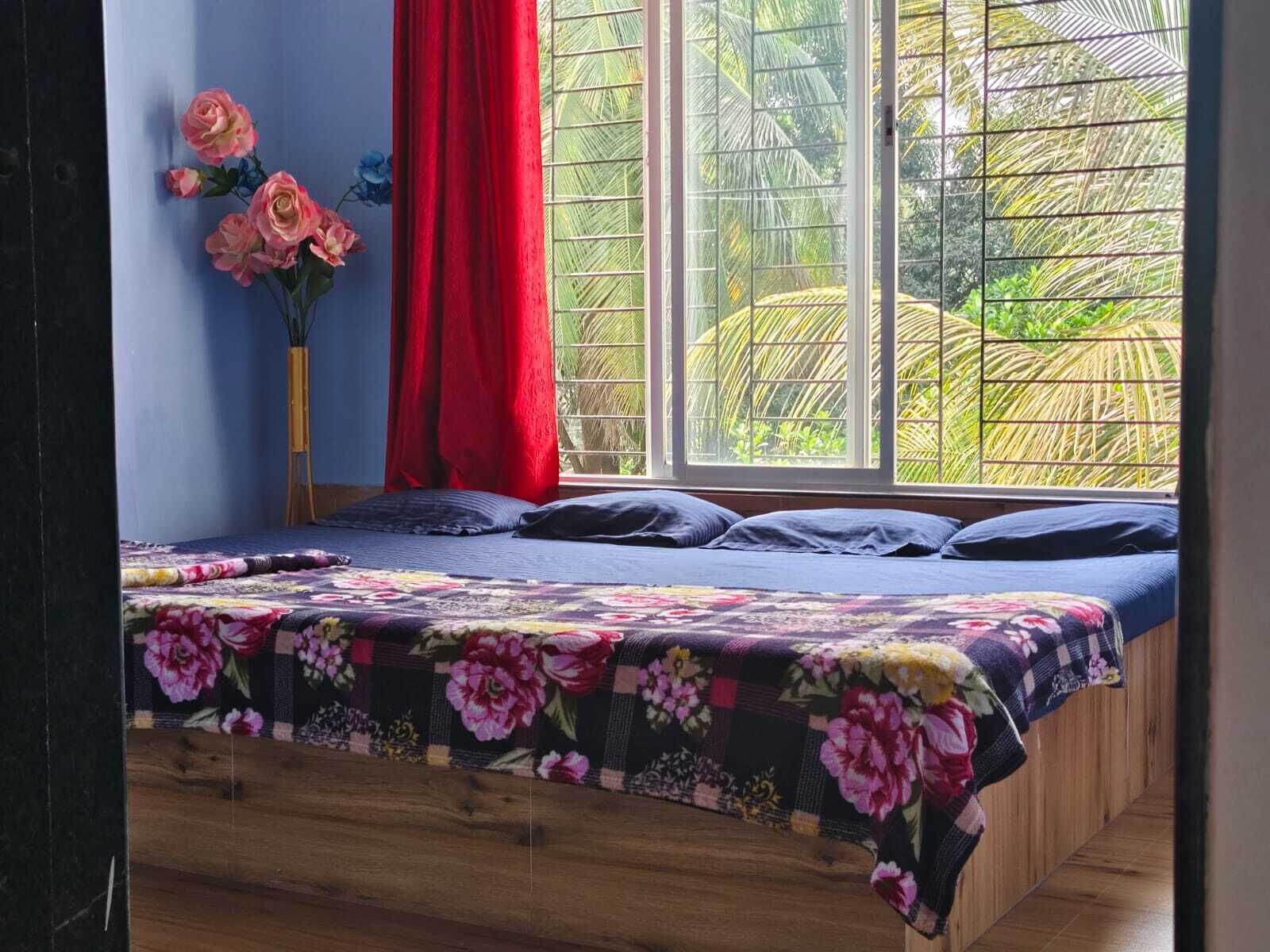 A bedroom with a wooden bed, floral bedding, a red curtain, and a view of palm trees through a window.