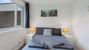 A bedroom with a bed, two bedside tables, a window with a view of a brick building, and a painting on the wall. - Spacious 4 Bedroom House With Parking (Gateshead)