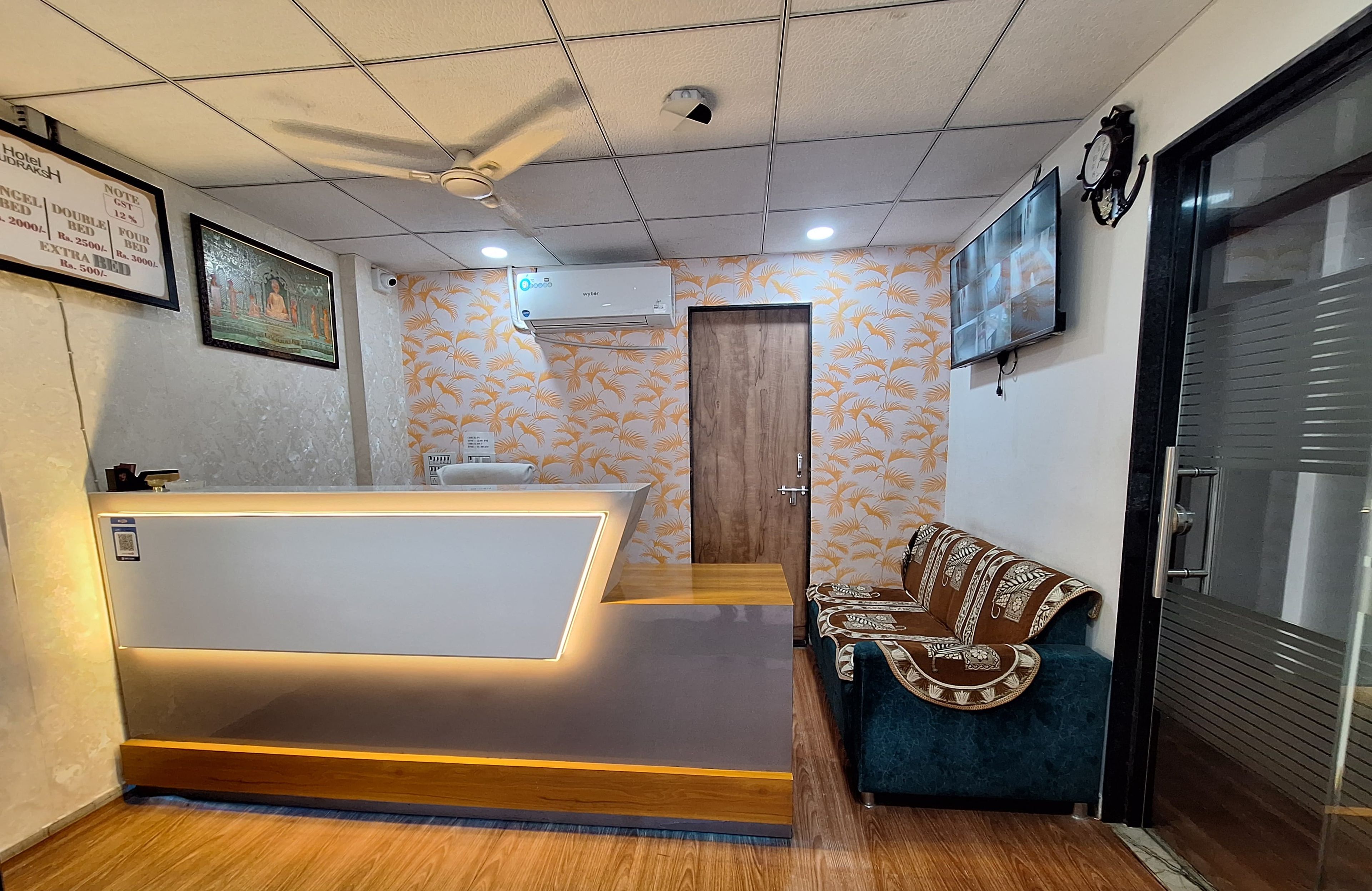 Zona de recepción de hotel con mostrador, un sofá, un reloj y un televisor montado en la pared.