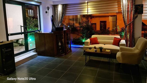 A modern living room with a sofa, armchair, and coffee table. There is a large window with curtains, a plant, and a view of the outside. - Paperon Butik Otel (Erdemli)