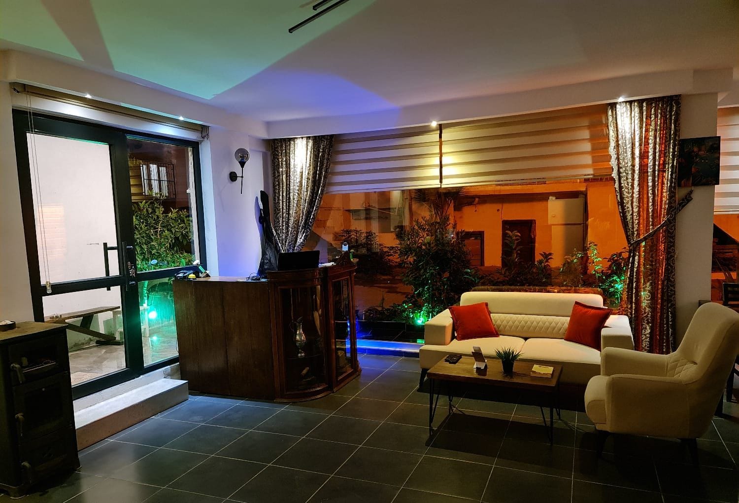 A modern living room with a sofa, armchair, and coffee table. There is a large window with curtains, a plant, and a view of the outside.