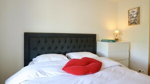 A bed with a dark headboard, white bedding, and a red pillow. There is a bedside table with a lamp and a framed quote on the wall.