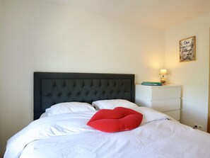 A bed with a dark headboard, white bedding, and a red pillow. There is a bedside table with a lamp and a framed quote on the wall.