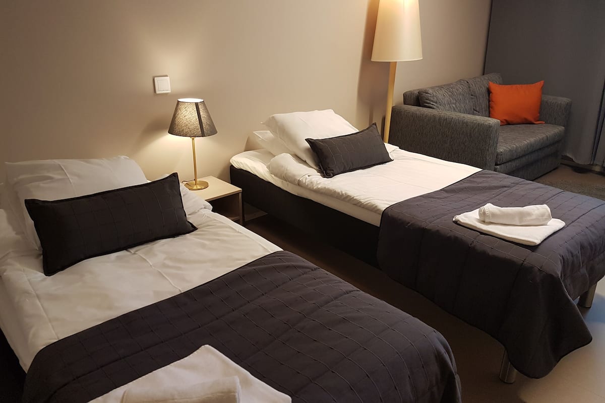 Een hotelkamer met twee bedden, een nachtkastje met een lamp, een grijze bank met een oranje kussen, en een groot raam.