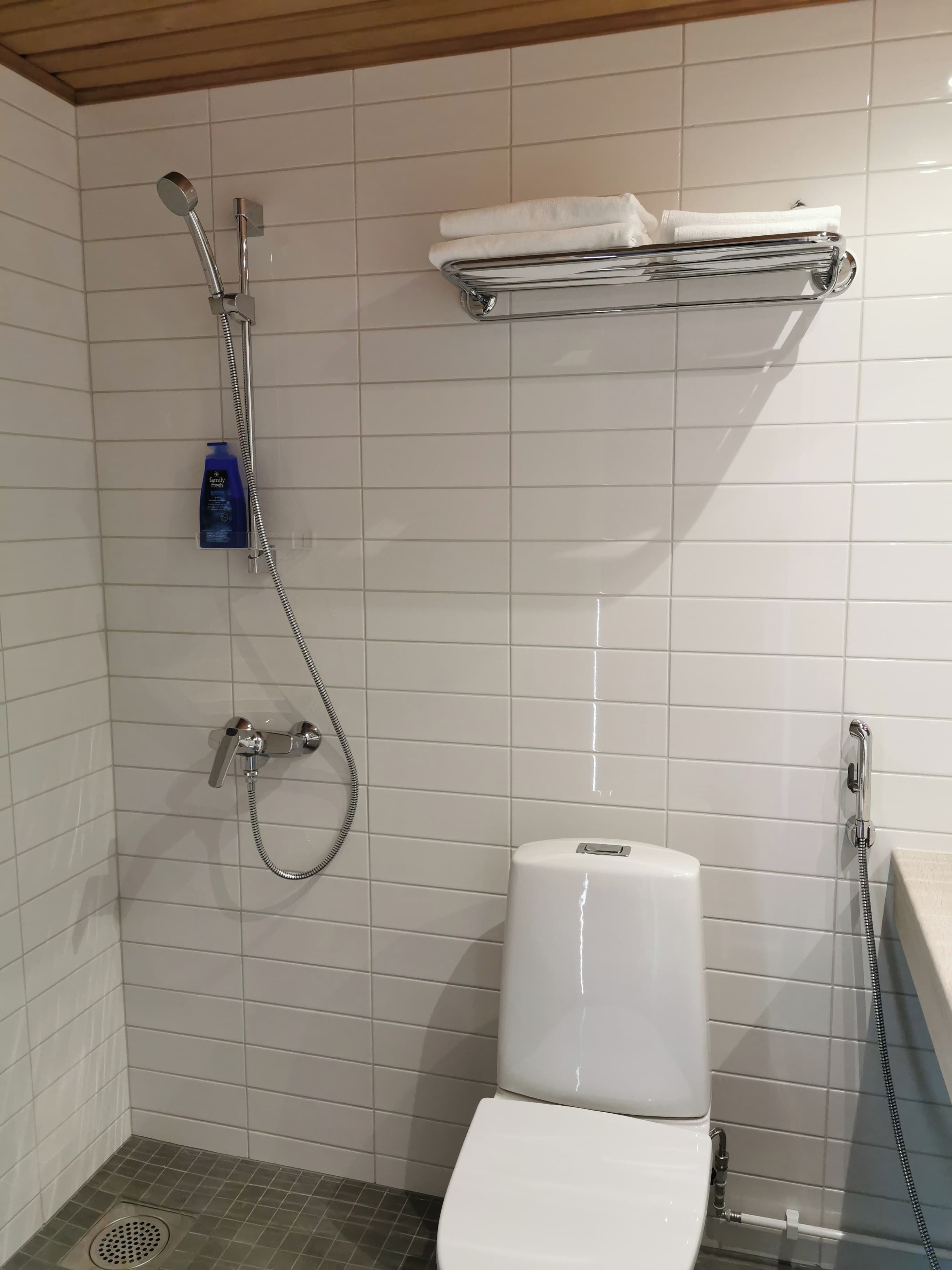 Um banheiro com paredes em azulejo branco, chuveiro com um chuveiro manual, vaso sanitário e uma prateleira com toalha.