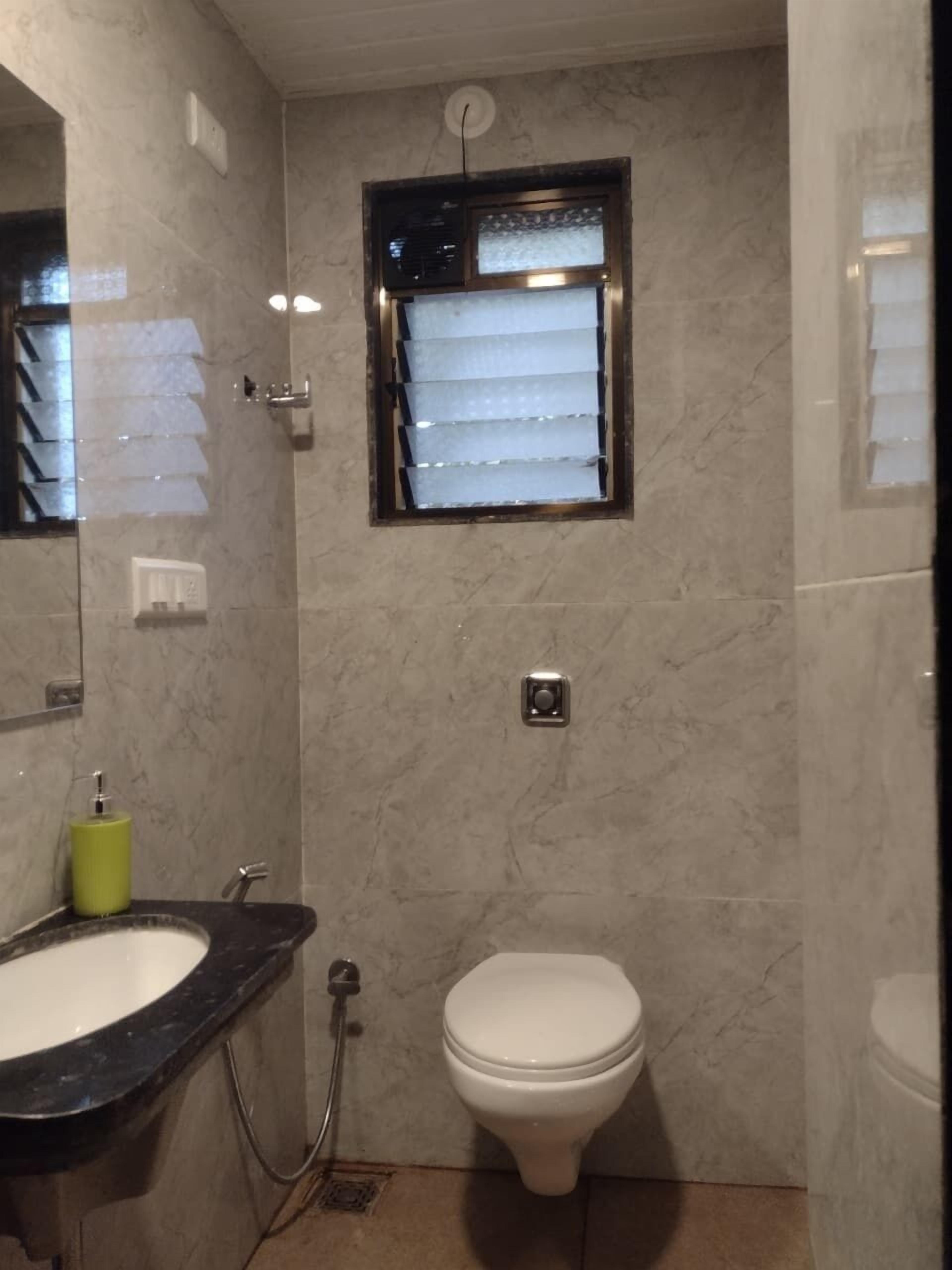 Ein Badezimmer mit einer Marmorwand, einer weißen Toilette, einem Waschbecken mit grünem Seifenspender und einem Fenster mit Jalousien.