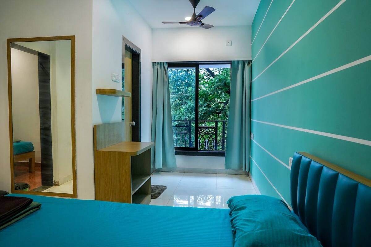 A bedroom with a bed, a desk, a chair, a window with a view of greenery, and a ceiling fan.
