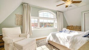 Un dormitorio con una cama, una silla, un aparador, una ventana con cortinas y un ventilador de techo.