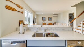 Una cocina moderna con un lavamanos doble, lavavajillas y horno empotrado. Hay paletas de madera en la pared, un ventanal grande y una escalera que sube a otro nivel.