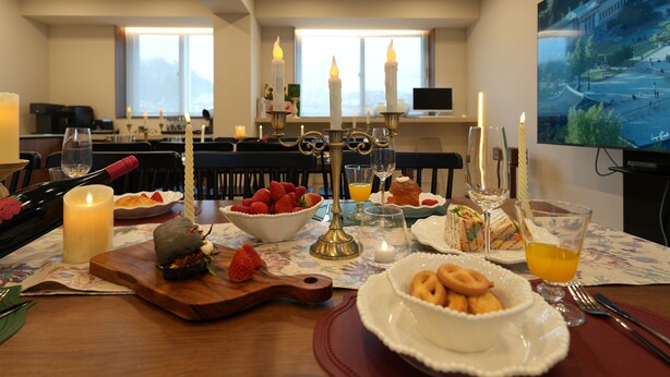 Un comedor con una mesa dispuesta para el desayuno, que incluye diversos alimentos y bebidas, velas y un televisor que muestra una vista panorámica.