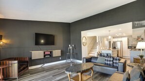 Una sala de estar moderna con un televisor de pantalla plana, una mesa de centro, sofás y una escalera que conduce a una cocina.