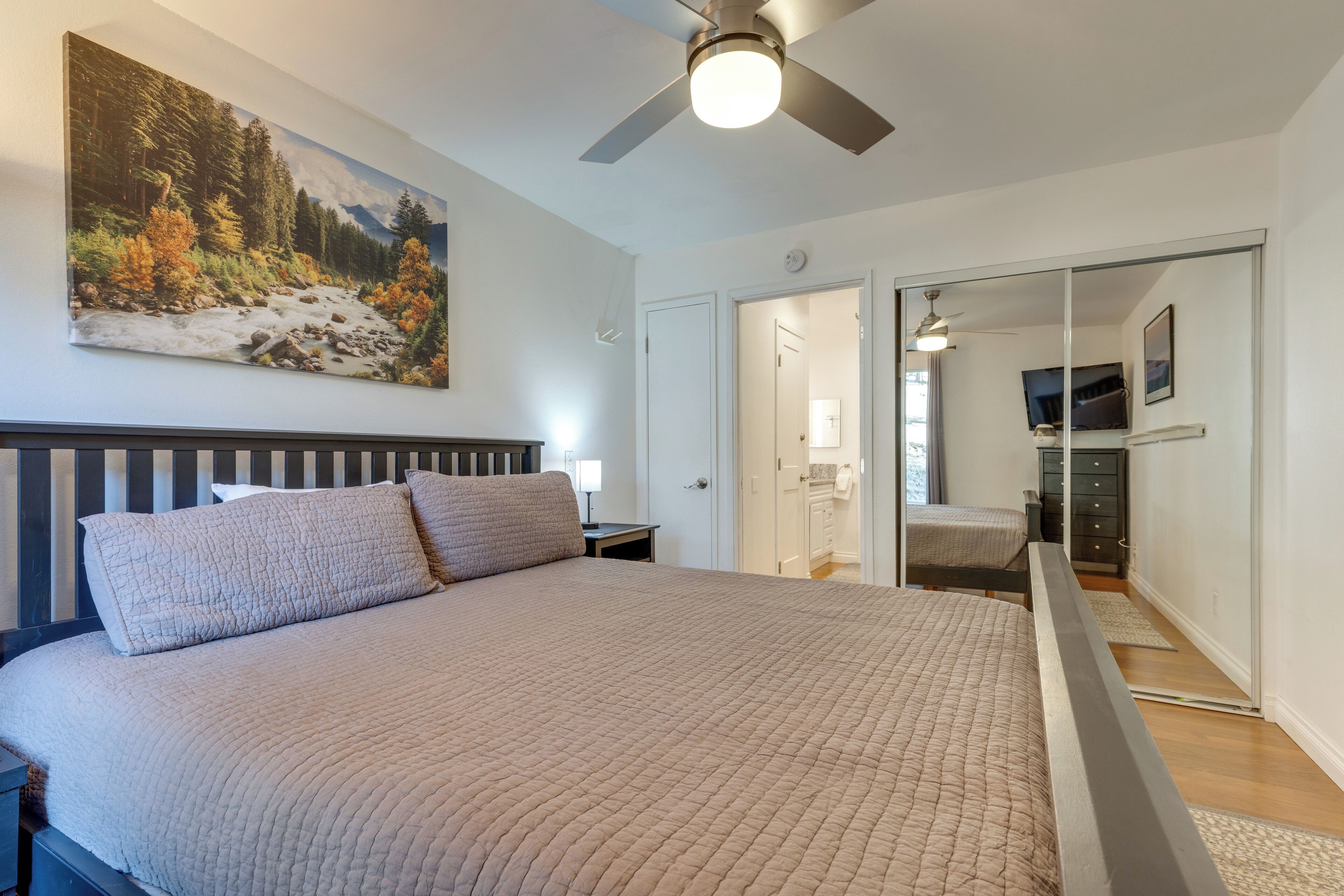 A bedroom with a large bed, a ceiling fan, a painting of a forest, and a sliding door leading to a bathroom.