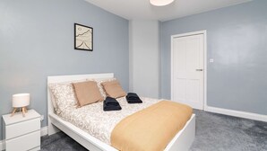 A bedroom with a bed, a nightstand, a lamp, a door, and a wall-mounted picture. - Spacious 3 BED House With Parking (Bo'ness)