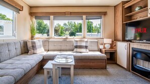 A modern living room with a sectional sofa, a fireplace, a TV, and large windows. - 3 Bed Caravan - Seton Sands (Prestonpans)