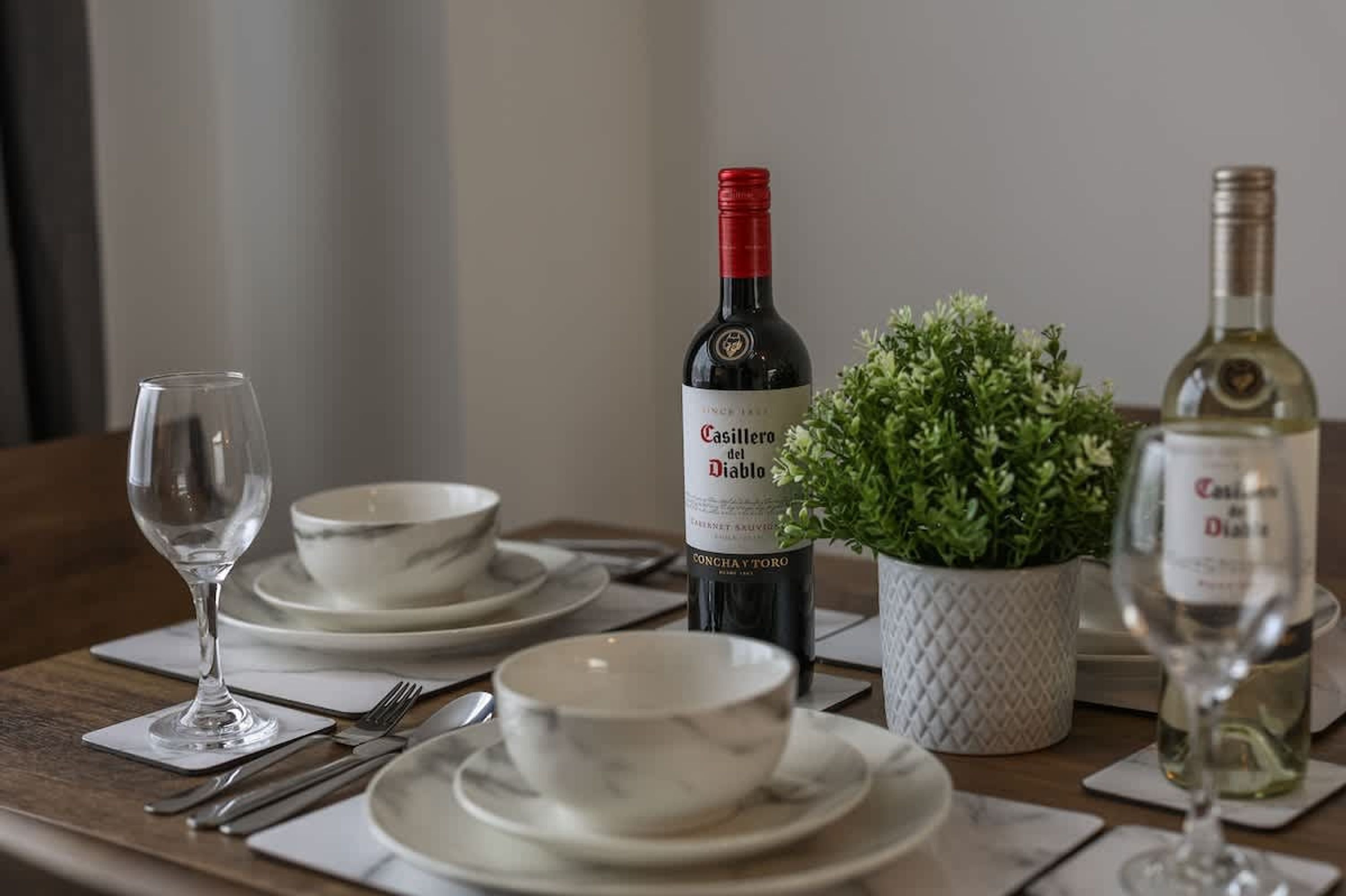 Una mesa de comedor preparada con platos, tazones, cubiertos y dos botellas de vino.