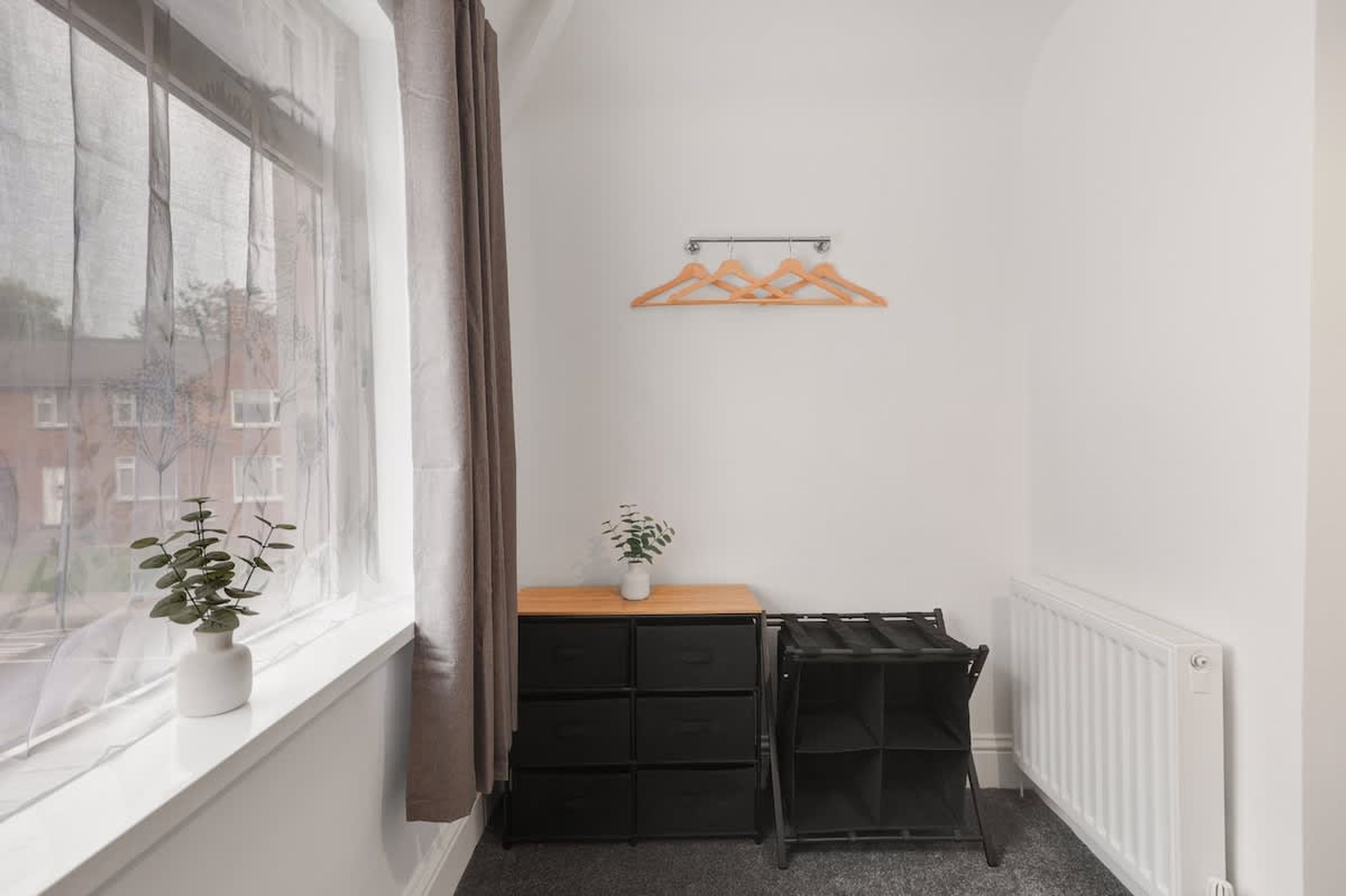 Een smalle kamer met een raam, een radiator, een zwart opbergmeubel en een houten plank met een plant.