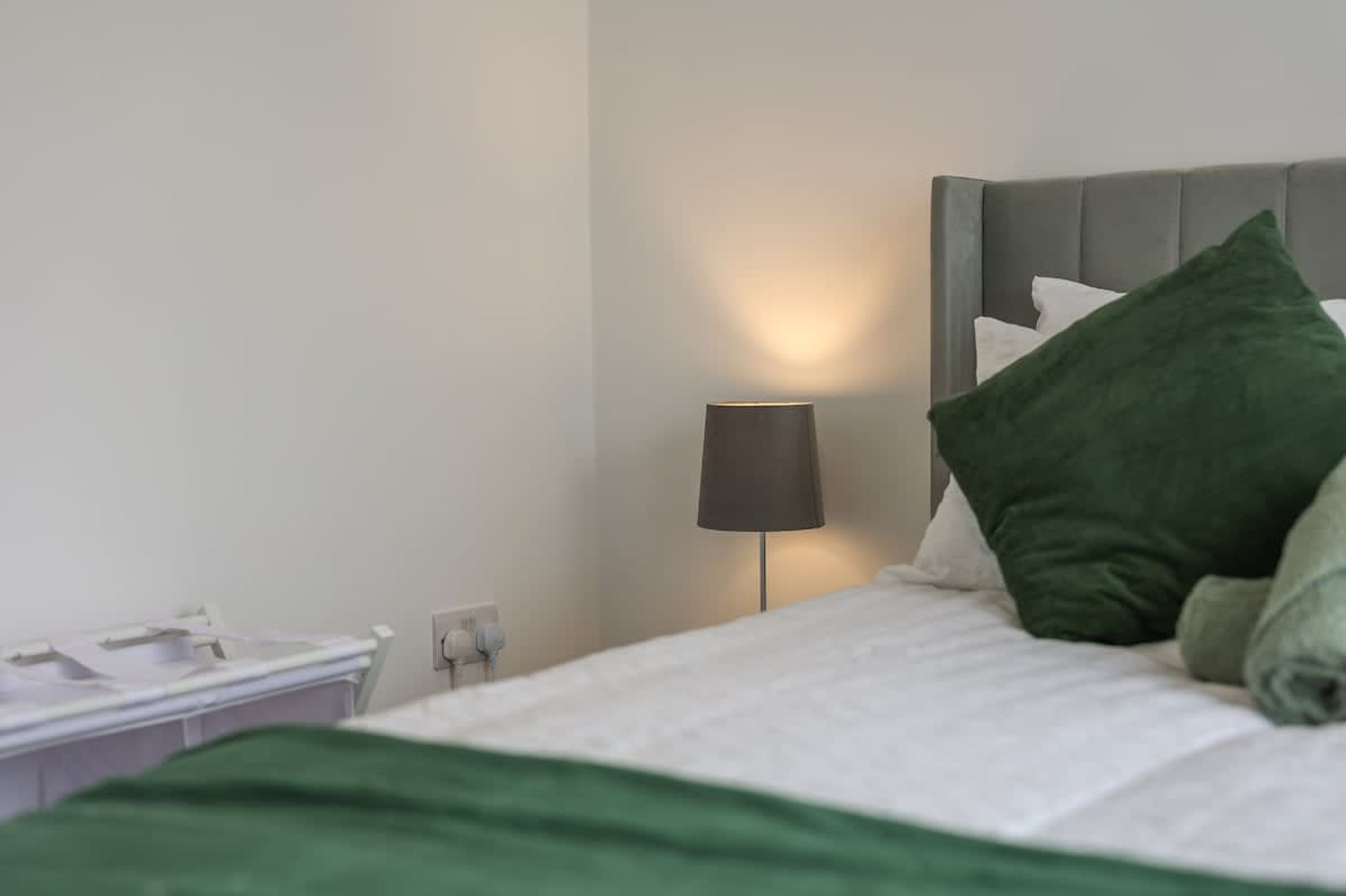 En pænt redt seng med en grøn pude, en senget lampe og et hvidt natbord.
