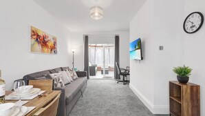 A modern living room with a grey sofa, a dining table set, a TV mounted on the wall, and a staircase leading to another room. - 3 Bedroom House With Free Parking (Durham)
