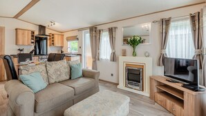 A living room with a sofa, ottoman, TV stand, fireplace, and kitchen area with cabinets and a stove. - Superb and spacious lodge with one double, one twin and one single. (Bala,)