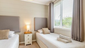 Ein Hotelzimmer mit zwei Betten, einem Nachttisch mit Lampe, einem Fenster mit Jalousien und Blick auf Grünflächen.