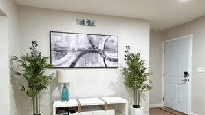 Un soggiorno moderno con un tavolino consolle bianco, due piante in vaso, un'opera d'arte con una veduta urbana incorniciata e una porta sulla destra.