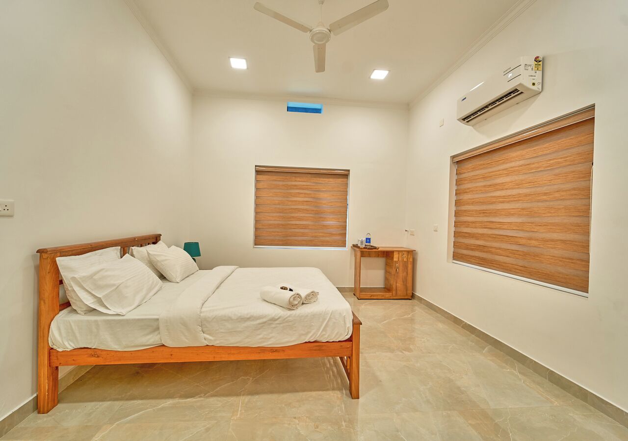 Une chambre à coucher avec un lit en bois, une table de chevet, un climatiseur fixé au mur et deux fenêtres équipées de stores.